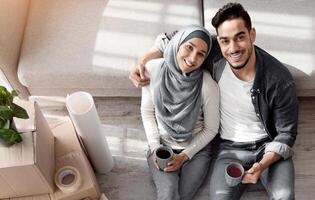 Loving arab husband and wife in hijab sitting on floor by couch in living room, having break while unpacking stuff at new house, hugging and drinking coffee, view above. Moving, recolating concept photo