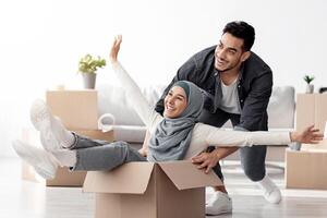 Emotional middle-eastern young family having fun while unpacking stuff in their new house. Happy arab guy and excited lady in hijab sitting in paper box, enjoying moving. Apartment, house, real estate photo