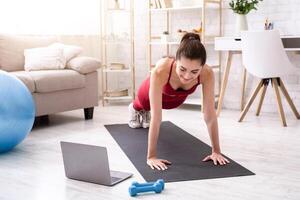 Personal trainer giving online workout class via laptop at home, doing plank or push up exercise photo