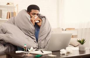 Sick black student working from home on laptop with homework assignment or university project, wrapped under blanket on couch, free space photo