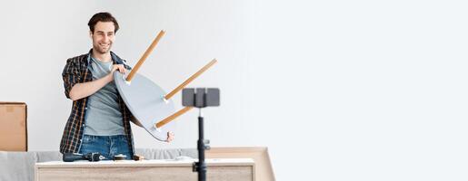 Remote work, blog about carpentry, furniture repair and assembly. Smiling millennial man with wireless headphones looks at phone in webcam and shows finished chair, in living room interior with tools photo