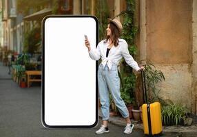 A woman stands in a lively street with a suitcase, holding her smartphone. She smiles next to a large, blank screen, surrounded by greenery and quaint buildings, suggesting an urban adventure. photo