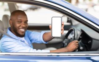 Closeup of young smiling African American man showing mobile phone with white empty screen for mock up sitting on driver seat in new luxury car, holding gadget in hand, selective focus on device photo