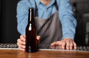 Barman in apron rests on wooden bar counter and serves bottle of beer without label, close up, cropped photo