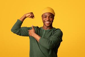 Cheerful Young Black Man Pumping Fist And Pointing At His Biceps, Positive African Millennial Hipster Guy Showing Muscles, Demonstrating Power And Strength While Posing Over Yellow Studio Background photo