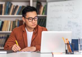 E-Teaching. Korean Middle Aged Man Teacher Using Laptop And Taking Notes Preparing For Virtual Lecture Sitting At Desk In Modern Classroom. Internet Technology And Education. Selective Focus photo
