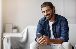 Handsome young indian guy sitting with smartphone in hands at home, messaging with friends, relaxing with cellphone on sofa, checking new app, enjoying mobile communication or browsing social networks photo