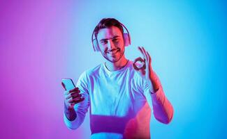 Symbol with hands is excellent. Smiling millennial guy with large wireless headphones expresses joy and holds smartphone, free space photo