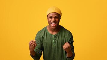 Hooray. Euphoric Excited Black Hipster Guy Celebrating Success With Raised Fists, Overjoyed African American Man Exclaiming With Excitement While Standing Isolated Over Yellow Background, Copy Space photo