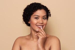 A woman with curly hair poses confidently, showing a joyful expression while gently touching her lips. The neutral background highlights her natural beauty and charm. photo