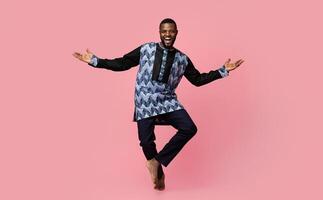 Full size photo of friendly black man dancing over pink studio background, copy space. Cheerful african american guy wearing traditional costume and showing welcoming gesture, studio shot