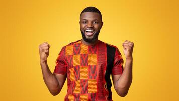 African man in traditional colorful clothes expressing happiness over yellow background, screaming and clenching fists photo