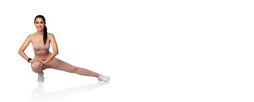 A young woman stretches her body in a graceful pose while wearing activewear. She smiles and focuses on her form, showcasing a dedication to fitness in a bright, minimalistic indoor space. photo