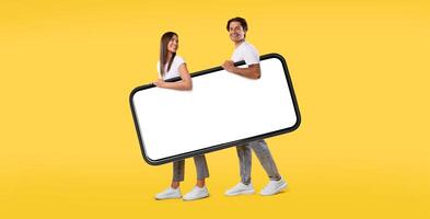 Cool Mobile App. Cheerful young couple holding big giant smartphone with mockup, walking together, promoting application or website, advertising your service or product over orange studio background photo