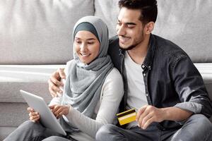 Smiling young muslim family husband and wife buying stuff for their new house, sitting on floor by couch among big paper boxes with belongings, using digital tablet and credit card, copy space photo