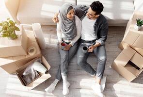 Top view of loving arab husband and wife in hijab sitting on floor by couch in living room, having break while unpacking stuff at new house, hugging and drinking coffee. Moving, recolating concept photo