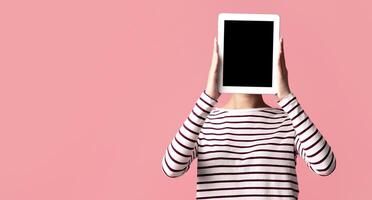 Unrecognizable Young Female Covering Face With Digital Tablet With Black Blank Screen, Woman In Striped T-Shirt Standing Over Pink Studio Background, Mockup Image With Copy Space For Your Design photo