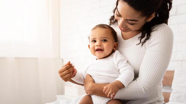 alegre africano americano madre abrazando bebé jugando con encantador niñito chico posando sentado en cama en dormitorio interior. niño cuidado y alegría de siendo mamá concepto foto