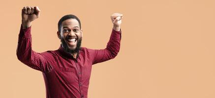 Winner Concept. Studio portrait of african american man yelling with clenched fists raised up in the air, isolated over yellow studio background. Black guy cheering and screaming, celebrating victory photo