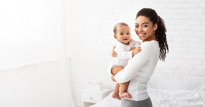 joven negro mamá participación bebé niño pequeño, abrazando que lleva adorable pequeño hijo posando en pie adentro. niño cuidado, maternidad y maternidad salir concepto. panorama, vacío espacio para texto foto