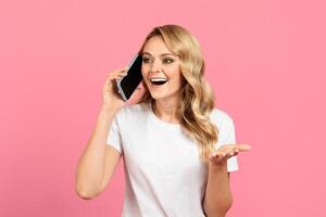 A young woman is engaged in an enthusiastic phone conversation, smiling brightly and gesturing with her hand. The backdrop features a vibrant pink color, enhancing her joyful expression. photo