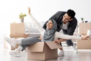Joyful middle-eastern young family having fun while unpacking stuff in their new house. Happy arab guy and excited lady in hijab sitting in paper box, enjoying moving. Apartment, house, real estate photo