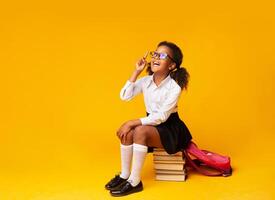 Idea Or Question. African American Schoolgirl Pointing Finger Up Sitting On Book Stack Over Yellow Studio Background. Copy Space photo