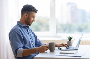 Remote Work. Handsome Male Freelancer Using Laptop And Drinking Coffee, Working Remotely At Home Office, Sitting At Desk Near Window, Browsing Internet, Having Online Job, Side View With Copy Space photo