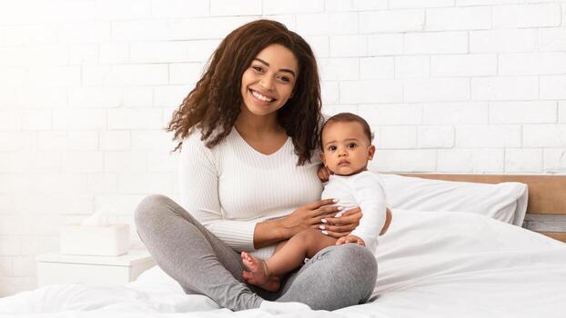 contento africano americano madre abrazando participación su infantil bebé chico sonriente a cámara sentado en cama a hogar. alegría de maternidad. niño cuidado y maternidad salir concepto foto