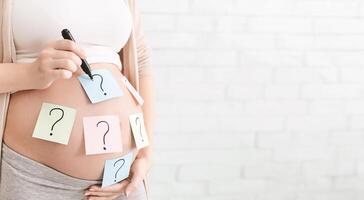 Concept of choosing baby name. Pregnant woman with question marks on paper stickers on tummy, panorama with empty space photo