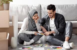 Excited arab couple working on design for their new apartment. Happy handsome middle-eastern man and woman in hijab sitting on floor among paper boxes with color samples, copy space photo