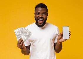 Making money on internet. Happy african-american guy showing showing cellphone with blank screen and bunch of money, orange background, free space photo