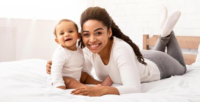 contento africano americano mamá abrazando bebé chico sentado en cama en dormitorio a hogar. negro madre posando con adorable niñito infantil sonriente a cámara adentro. alegría de maternidad, niño cuidado concepto foto