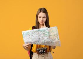 Confused woman traveler with camera looking at city map, got lost, yellow studio background, copy space. Upset young lady backpacker lost in city, checking map. Navigation, trekking during travelling photo