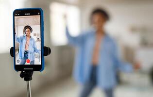 Modern Content Creator. Positive African American young lady in headset filming blog for social network, using cellphone on tripod for broadcast in living room, selective focus on device screen photo