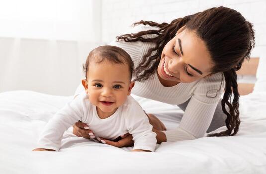 africano madre jugando con linda bebé hija Ayudar su gatear en cama en dormitorio a hogar. joven mamá disfrutando maternidad rutina y niño cuidado. maternidad salir estilo de vida foto