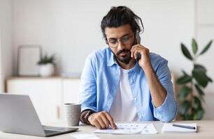 Young Arab Male Entrepreneur Talking On Cellphone And Working With Papers In Modern Office, Handsome Eastern Guy Sitting At Desk Checking Financial Reports And Speaking On Mobile Phone, Free Space photo