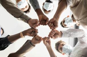 Success And Teamwork Concept. Below view of young diverse group of business people in protective medical masks making fist bump standing in circle. Workers do fist pump together celebrating good deal photo