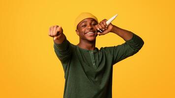 Joyful young black man singing with microphone and pointing at camera, funky cool african american hipster guy having fun, millennial mc standing isolated over yellow background, copy space photo