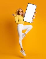 Cheerful blonde woman jumping up and showing newest smartphone with empty screen, enjoying new application for mobile device. Orange studio background, creative collage with mockup photo
