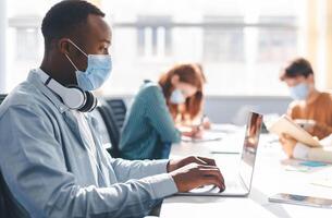 Group of diverse people sitting at the table, working, studying for test, making project or homework together, wearing surgical masks. African american man with headphones using laptop at workplace photo
