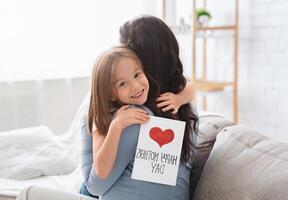 Smiling little daughter with Happy Mother's Day gift card hugging her mom, celebrating International Mother's Day together at home photo