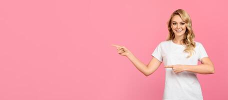 A young woman with long hair and a cheerful expression stands in front of a vibrant pink wall. She is pointing to blank space with both hands, creating an inviting and playful atmosphere. photo
