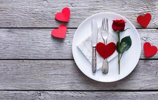 Romantic dinner concept. Valentine day or proposal background. Top view of restaurant wooden table with heart and rose with cutlery on plate. Copy space on rustic wood photo