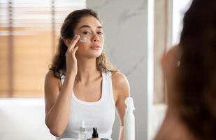 mujer aplicando facial hidratante crema en cara conmovedor las mejillas en baño a hogar. hembra protección de la piel rutina y productos cosméticos, belleza y mimos concepto. selectivo atención foto