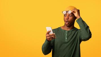 Amazing App. Shocked African American Guy Looking At Smartphone Screen, Amazed Black Millennial Man Taking Off Eyeglasses While Reading Unexpected Message, Standing Over Yellow Background, Copy Space photo