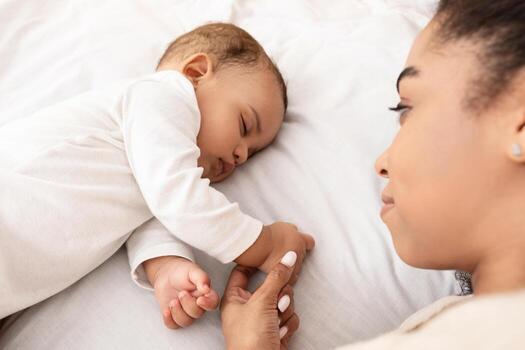 vista desde arriba de dormido africano americano bebé infantil y su madre acostado en cama en dormitorio a hogar. mamá participación su pequeño niñito hijos mano mientras él duerme pacíficamente selectivo atención foto