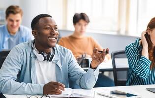 Learning Concept. Portrait of interested African American guy sitting at table with multi ethnic classmates, talking to teacher, asking or answering question, discussing new theme, topic or theory photo