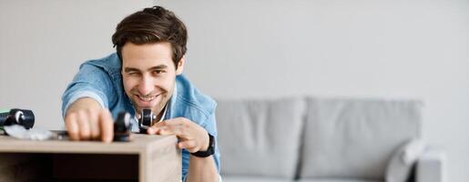 Self made modern furniture and hobby, engineer with tools, homemade decoration. Young smiling man assembling wooden bookshelf or desk measures distance between wheels on table in living room interior photo