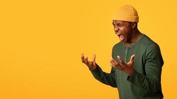 Portrait Of Angry African American Man Screaming With Rage, Furious Agressive Young Black Guy Raising Hands And Shouting While Standing Over Yellow Background In Studio, Panorama With Copy Space photo